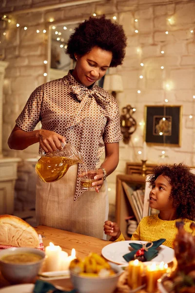 Happy African American mother serving juice to her daughter while during a meal on Thanksgiving at dining room.