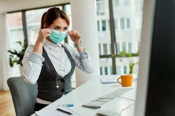 Young businesswoman using computer and putting on protective mask on her face in the office.