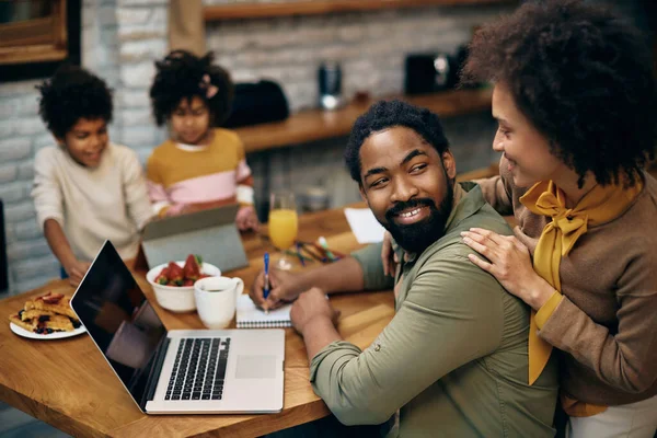 Happy African American couple talking while using computer at home. Their kids are using touchpad in the background. Focus is on man.