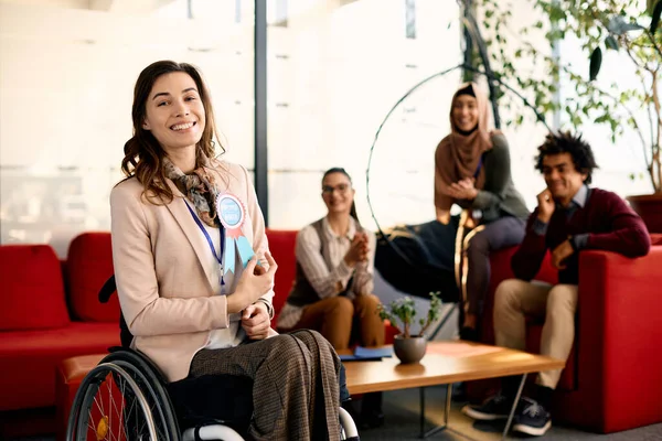 Happy businesswoman in wheelchair receiving employee of the month badge in the office and looking at camera. Her coworkers are in the background.