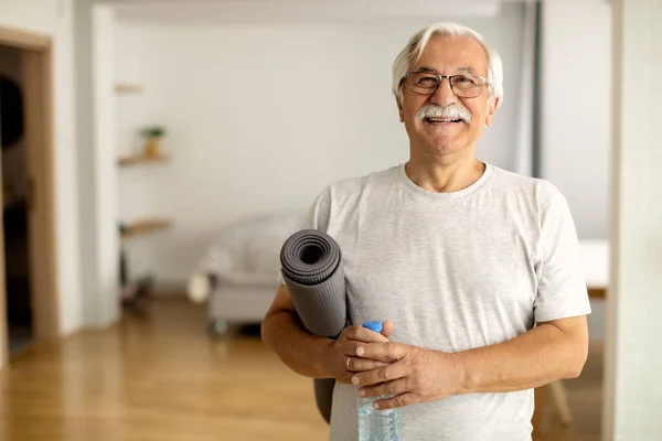 Happy senior man holding bottle of water and exercise mat in the living room and looking at camera.