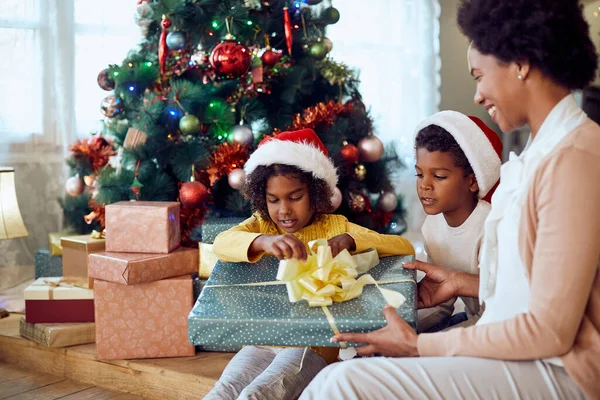 Happy African American family enjoying while opening gifts by Christmas tree at home. Focus is on kids.