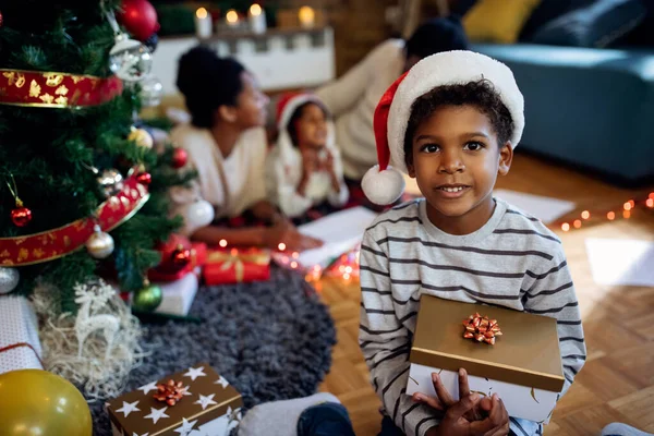 Small African American boy holding Christmas present while wearing Santa hat at home. His family is in the background.
