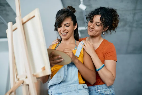 Young gay woman painting on canvas while her girlfriend is embracing her at home.