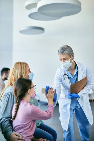 Happy female doctor and little girl greeting with high-five gesture in waiting room at the hospital. They are wearing face masks due to COVID-19 pandemic.