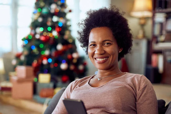 Happy black woman using smart phone while relaxing at home for Christmas and looking at camera.