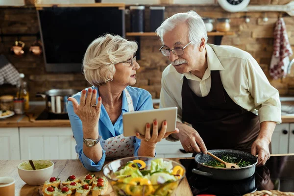 Senior couple communicating and following recipe on digital tablet while preparing food in the kitchen.