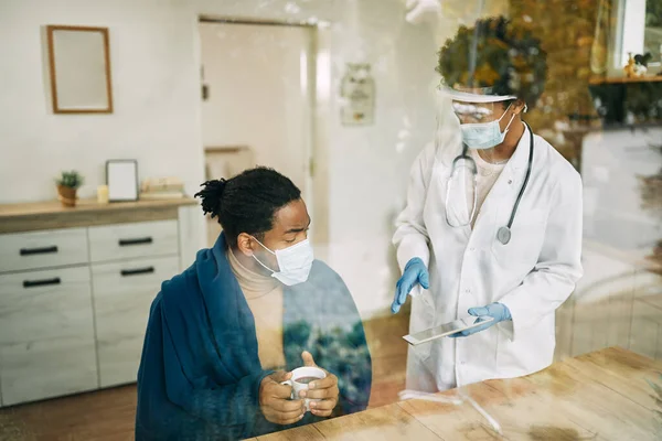 Black man and his female doctor talking while using digital tablet and wearing face masks during home visit. The view is through the glass.