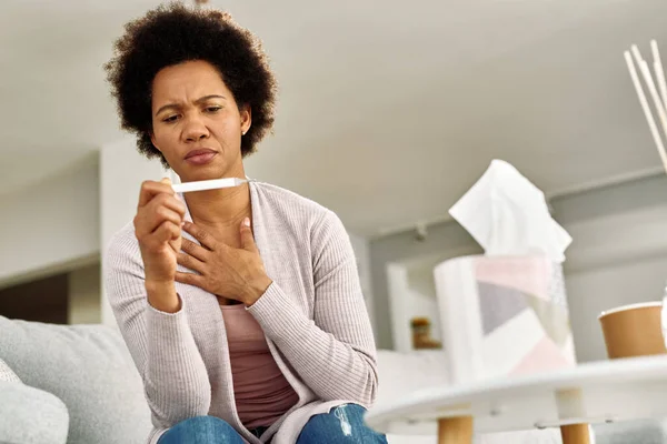 Low angle view of black woman having chest pain and measuring temperature at home.