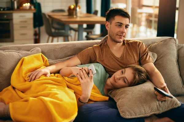 Young man watching TV while relaxing on the sofa with his girlfriend who is using smart phone. 