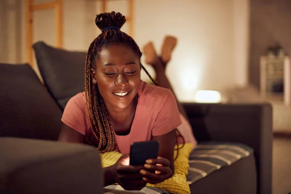 Happy black woman using smart phone and text messaging while relaxing at night in the living room.