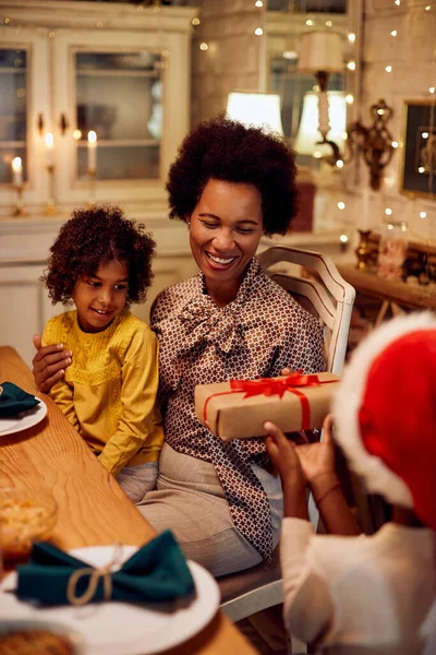 Happy black mother enjoying in Christmas while receiving a gift from her son and daughter at home.