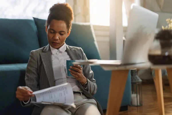 Black female entrepreneur drinking coffee and analyzing business reports while working at home.