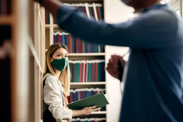 College student wearing face mask and talking to her friend while studying in a library.