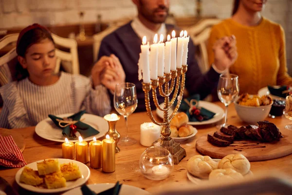 Close-up of dining table with lighted candles in menorah at dining table on Hanukkah. Family in the background is holding hands and praying.