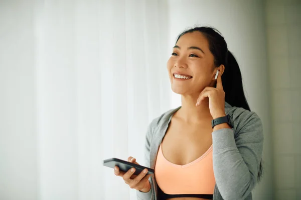 Young Asian athletic woman enjoying in music over earphones while using mobile phone in dressing room at the gym.