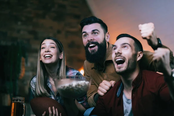 Small group of young excited people watching sports match on TV at home.