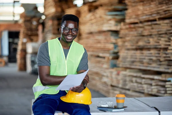 Happy black warehouse worker analyzing plans at lumber distribution department and looking at camera.