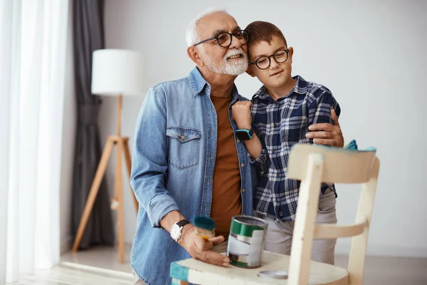 Loving senior man embracing his grandson while spending time with him and painting furniture together at home.