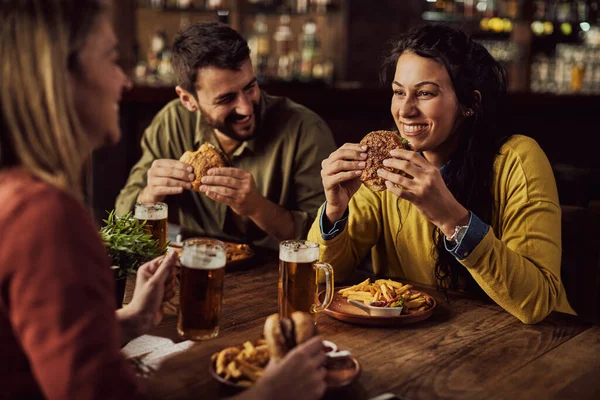 Happy woman having fun while talking to her friends and eating hamburger in a pub.