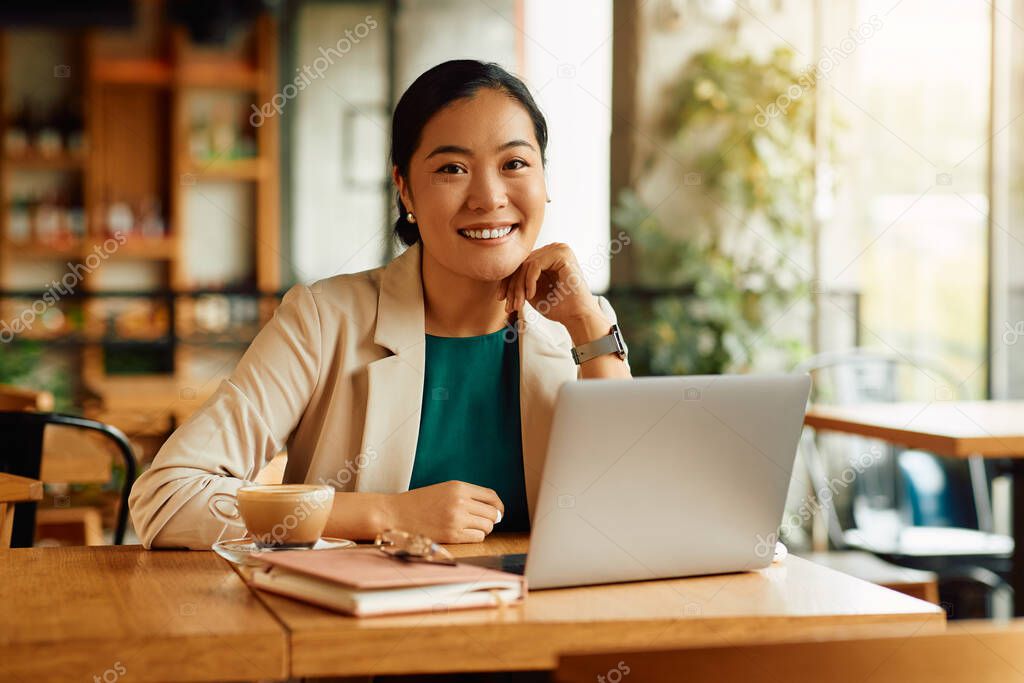 Portrait of Asian female entrepreneur working on laptop while drinking coffee in a cafe and looking at camera.