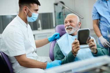 Satisfied senior man looking his teeth in a mirror and talking to his orthodontist after dental appointment at dentist's office. 