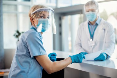 Nurse wearing protective mask and face shield while working at reception desk at medical clinic during coronavirus pandemic. 