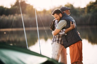 Happy man embracing his senior father while fishing with his during their camping day by the lake. 