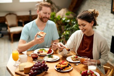 Happy couple having a breakfast in the morning at home. Focus is on woman pouring honey on waffles. 
