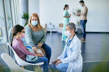 Küçük hastasıyla konuşan çocuk doktoruyla ilgilenen doktor annesiyle birlikte hastaneye geldi. COVID-19 salgınından dolayı koruyucu yüz maskeleri takıyorlar..