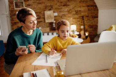 Small girl using laptop and writing on the paper while mother is asissting her at home. 
