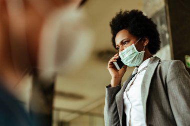 Koruyucu maskeli siyahi iş kadını tren istasyonu koridorundayken cep telefonuyla konuşuyor.. 