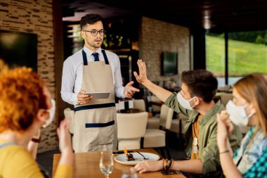 Group of customers gesturing stop sign to a waiter who is not wearing protective face mask in a cafe during coronavirus epidemic. Focus is on waiter. 