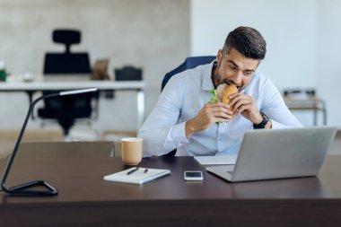Male entrepreneur reading an e-mail on a computer while eating sandwich at his office desk. 