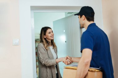 Young happy woman standing on a doorway and shaking hands with delivery man while receiving a package.