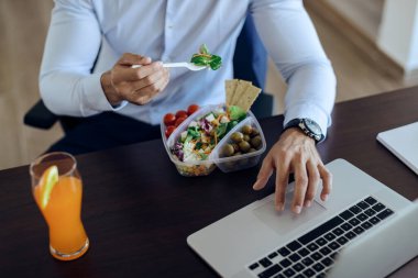 Unrecognizable businessman eating healthy food and surfing the net on laptop on lunch break in the office. 