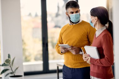 Two entrepreneurs communicating in the office and wearing protective face masks due to COVID-19 pandemic. Focus is on man using touchpad.