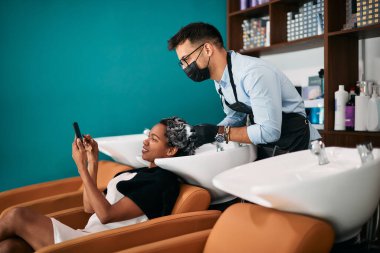 Happy African American woman showing something on cell phone to her hairstylist while he is washing her hair at the salon.