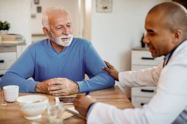 Senior man communicating with black doctor who is visiting him at nursing home. 