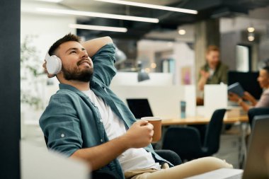 Young smiling entrepreneur relaxing and listening music over headphones while having coffee break at corporate office. 