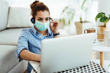 Female student with protective mask using laptop while e-learning from home during virus epidemic.   