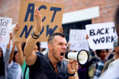 Young displeased activist using megaphone and shouting while protesting with crowd of people against government recession measures. 