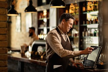 Happy waiter using cash register while checking orders and working in a pub. 
