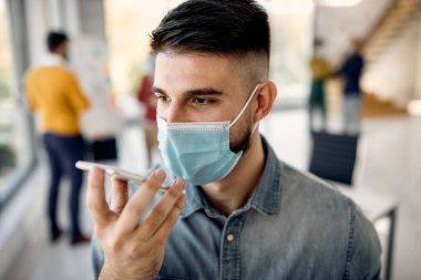 Male entrepreneur wearing protective face mask talking over mobile phone's speaker in the office.