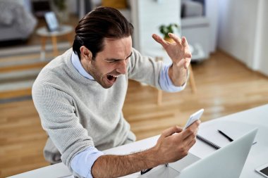 Angry businessman screaming from frustration while reading bad news over cell phone at home. 