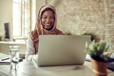 Happy Islamic businesswoman working on a computer and communicating over headset in the office. 