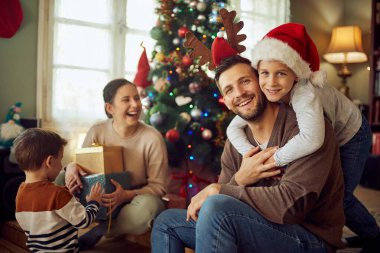 Happy parents with kids having fun while spending Christmas day together at home. Focus is on embraced father and son looking at camera. 