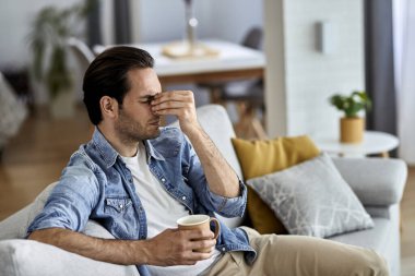 Young man having a headache while drinking coffee in the living room. 