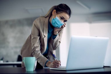 Businesswoman taking notes while using laptop and talking on the phone in her office. She is wearing face mask due to COVID-19 pandemic. 