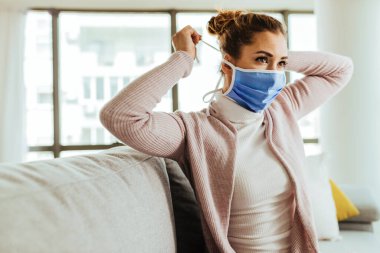 Young woman using protective face mask at home during coronavirus pandemic. 
